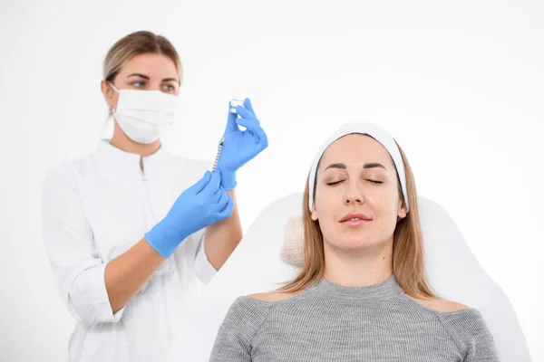 front view on female patient lying on couch in beauty clinic and female cosmetologist in blue latex gloves on background. Cosmetology concept.