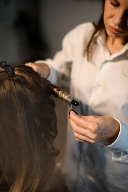 professional stylist curls long female hair with curling iron and makes styling for woman client.