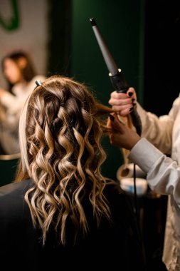 hairdo making process, stylist curls long female hair with modern clipless curling iron, close-up, hair salon