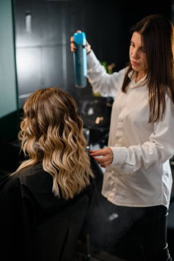 Selective focus on woman with long curling hair in hair salon and female hairdresser with hairspray bottle at background