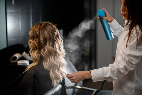 Beautiful young woman with long curly hair in hair salon and professional hairdresser styling her hairstyle with hairspray.