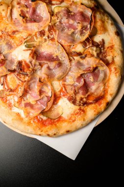 Cropped photo of round pizza with bacon, cheese and slices of pickled cucumber and onion on wooden chopping board on black table