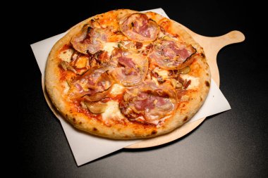 view on round pizza with bacon, cheese and slices of pickled cucumber and onion on wooden chopping board on black table