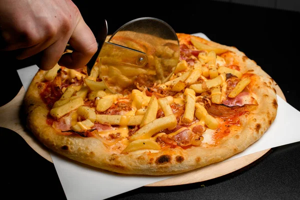 hand cuts with a pizza cutter delicious hot pizza with topping of french fries and bacon slices. Traditional italian cuisine