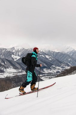 side view of male skier with ski equipment walking at snow against the backdrop of beautiful winter mountain landscape and sky