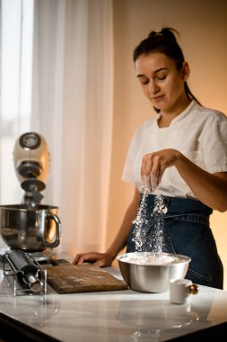 woman gently pouring by hand white flour in stainless bowl on table with modern kitchen appliances. Woman prepares dough pouring flour into steel bowl.