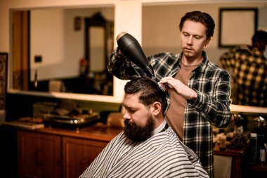 master barber does the hairstyle and styling with dryer, dries hair to bearded guy. Concept barbershop.
