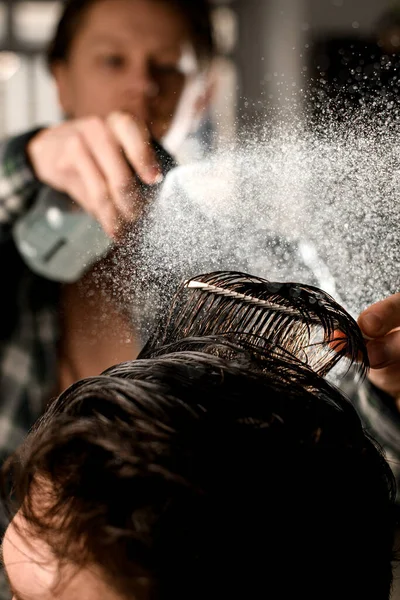 Close-up of head hair of male client which barber wets by spray and combs. Man barber making modern male hairstyle