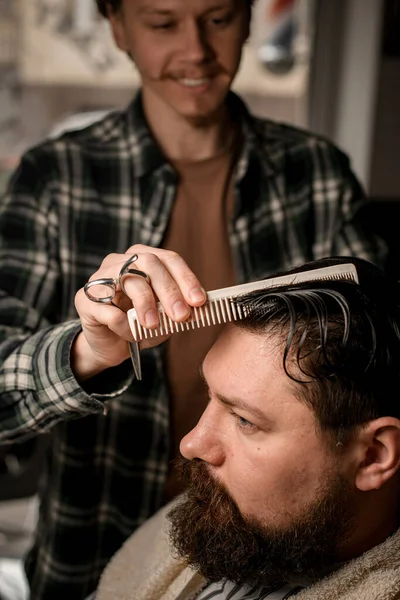 Close-up view of barber accurate combs wet male hair and make new style for client. Stylish bearded man getting trendy haircut and hairstyle. Concept of hairdressing and shave.