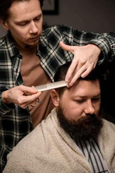Professional barber accurate comb wet male hair and make new style for his demanding client. Stylish bearded man getting trendy haircut and hairstyle. Concept of hairdressing and shave.