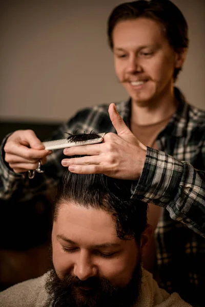 Selective focus on hands of professional barber cutting and styling hair with comb to his client. Bearded man getting trendy haircut and hairstyle. Barbershop concept