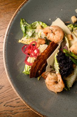 Kesilmiş fotoğraf. Tabağın üstünde karides ve kızarmış pastırma dilimleri ve masada taze marul var.