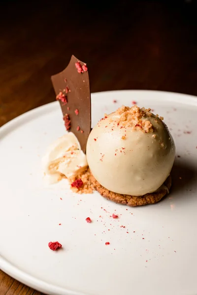 stock image great view on sweet dessert of white chocolate sphere garnished with biscuit crumble and ice cream with slice of dark chocolate on plate