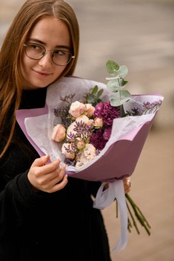 Nazikçe büyük ve güzel bir buket taze gül, okaliptüs, limonyum ve karanfilleri krema, menekşe ve bordo renklerinde tutan güzel bir kız.