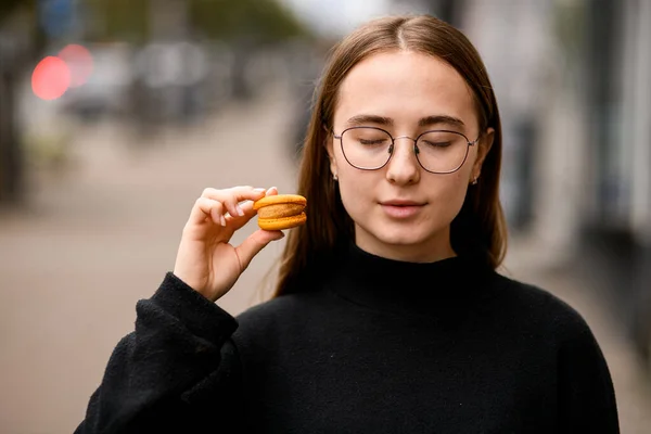 Portrait of beautiful girl with closed eyes holding orange macaroon in hand and dreaming of its rich taste