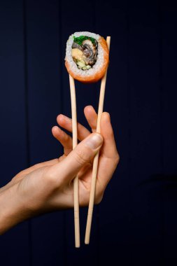 İştah açıcı Philadelphia dürümlerinden birinin yan görüntüsü. Elinde bambu çubuklarıyla koyu arkaplanda.
