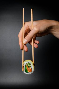 İnsanlar ellerinde ahşap çubuklarla Philadelphia suşi rulosu, füme somon, avokado, salatalık ve siyah arka planda yeşil buzağı tutuyorlar.