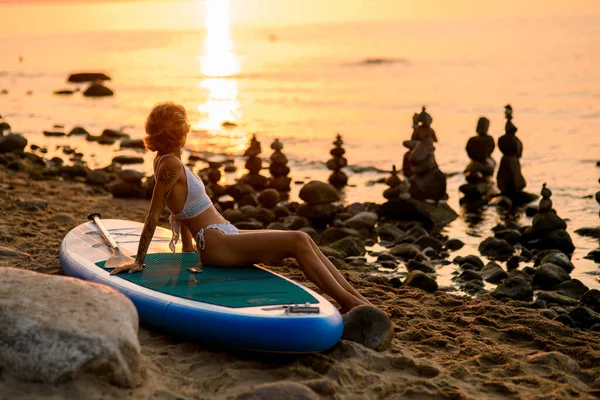 Young woman wearing white bikini sitting on the surf board for sup surfing on the beach at the sea on sunset of day, holliday summer vacation concept,shilouete photo,
