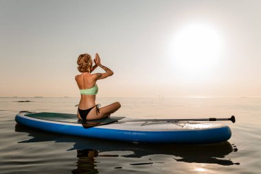 Nilüfer çiçeğinde oturan genç kadın, güneşin doğuşunda ya da batışında sessiz denizde sörf tahtasında yoga yapıyor.