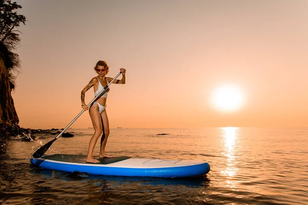 stock image Young tourist woman floating on the sup board over the sea. Amazing orange sunset and rock cliff on background