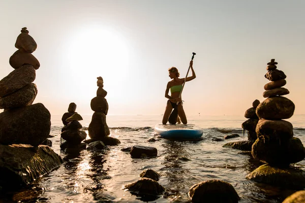 Pretty girl floating on sup surfing board between stone towers at sea during sunrise or sunset.