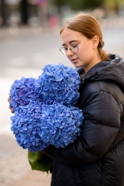 Çekici bir çiçekçi kız nazikçe el ele tutuşup büyük bir buket taze ortanca çiçeğine mavi renklerle bakıyor.