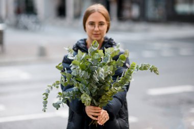 Genç çekici çiçekçi kız sokakta duruyor ve elinde taze yeşil okaliptüs dallarıyla bulanık arka planda duruyor.