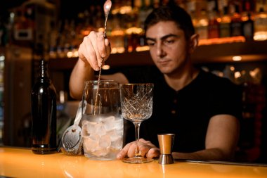 Barmen uzun bir kaşık tutuyor ve karıştırma kabına buz parçaları serpiyor. Kokteyl yapmak için araç gereç