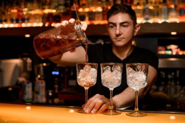 Bartender pouring an alcohol from the measuring cup through the strainer to cocktail glass with ice cubes on the bar counter in the blurred background clipart