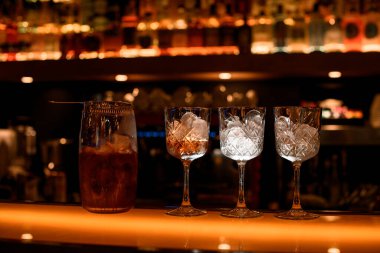 İçinde kokteyl olan şeffaf karıştırma bardağı olan bir tezgah. Yakınlarda üç kokteyl bardağı duruyor. Arka planda bulanık şişeler olan raflar.