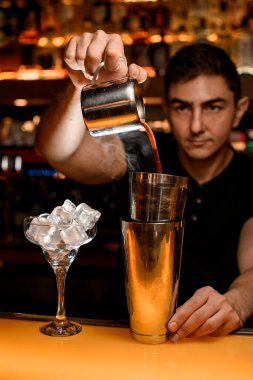 Barmen bakır bardaktan çelik bardaklara alkol dolduruyor. Yanında buz küpleri olan martini bardağı duruyor.