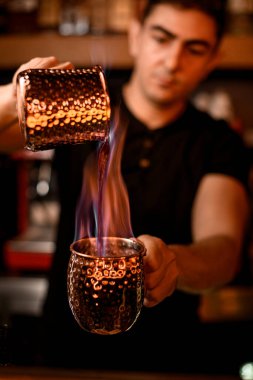 Yakışıklı barmen yanan alkolü bir bardaktan diğerine dolduruyor. Arka planda bulanık şişeler olan raflar