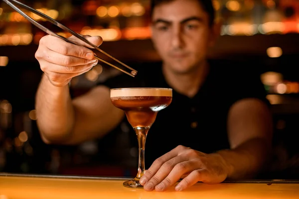 Closeup view on glass with espresso martini cocktail which bartender decorates with small coffee bean using tweezers