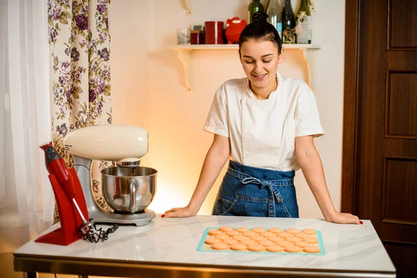 Woman stands in kitchen near food processor with metal bowl, smiles and looks on freshly baked halves of macaron dessert on silicone mat on kitchen table