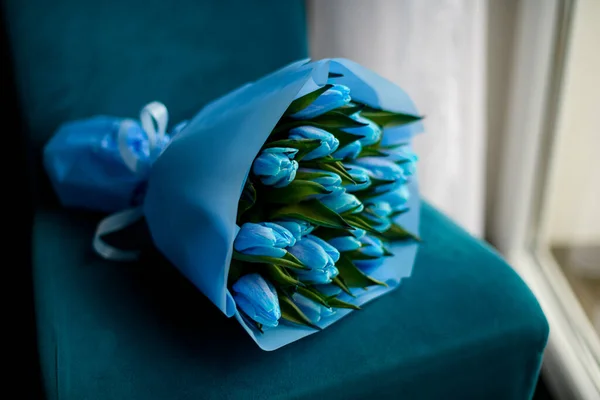 Side view of large bouquet of fresh blue tulips wrapped in blue packing paper on blue velvet chair. original bouquet of flowers for special event