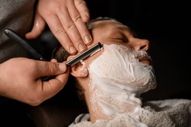 Profesyonel berber, modern berber dükkanında genç ve çekici müşteriyi klasik ustura ve paslanmaz çelikle tıraş ediyor. Yakından bak. Bulanık arkaplan