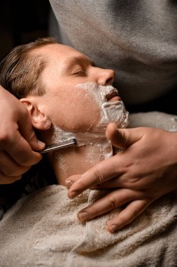 Modern berber dükkanında sandalyede otururken erkek berbere tıraş olan çekici bir genç. Kişisel bakım teması