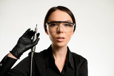 Siyah üniformalı, yüzünde koruyucu gözlüklerle manikür makinesi tutan kadın portresi. Beyaz arka plan. Metin için boşluk