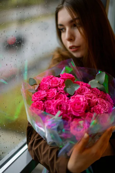 Beautiful brunette woman leans on window and holds large bouquet of colorful fresh roses wrapped in transparent packaging