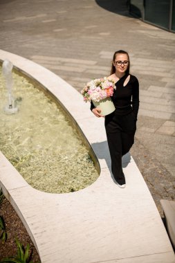 Çeşmenin yanındaki çekici kız elinde taze renkli çiçeklerle şapka kutusu tutuyor. Bulanık arkaplanda cadde