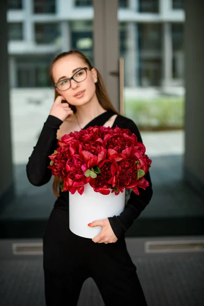 Beautiful brunette girl accurate holds in hands big round box with large dark red peony flowers. Blurred background. Flowers shop concept