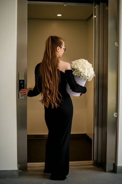 Stylish girl in total black look waiting for elevator and holding in hand flower bouquet of white hydrangea and gypsophila in hat box. Flover delivery concept. Gift box with beautiful flower