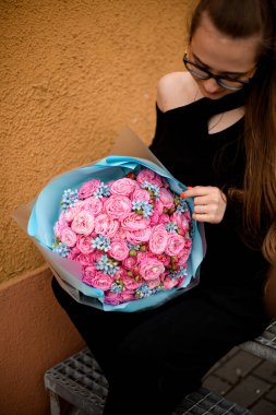 Güzel şık giyimli kız turuncu duvarın yanında oturuyor ve elinde mavi el sanatları kağıdında küçük mavi çiçeklerle süslenmiş büyük bir buket taze gül tutuyor. Bir demet çiçekli kadın. Üst görünüm