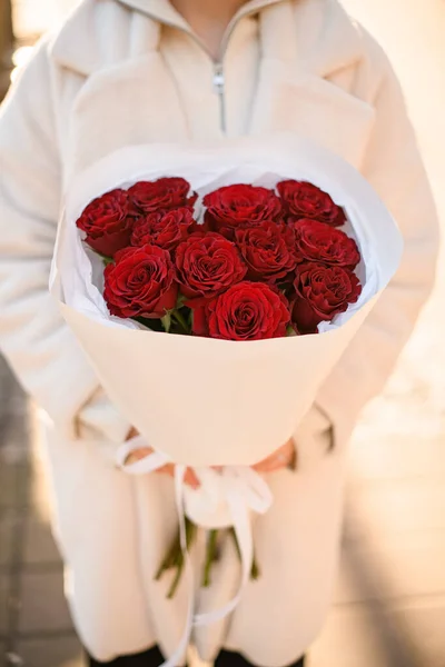 Selective focus on bouquet of bright red roses in white wrapping paper in womans hands. Flower shop concept. Vertical view