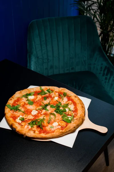 Appetizing Italian pizza with tomato sauce, arugula, soft cheese and chicken pieces on wooden board in restaraunt interior. Vertical orientation, top view