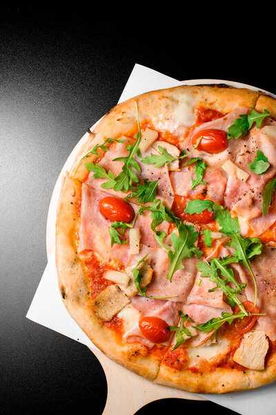 Fresh wood fired pizza with prosciutto di parma, arugula, halved cherry tomatoes, pineapple and parmesan on black background. Italian traditional cuisine. Delicious food. Top view