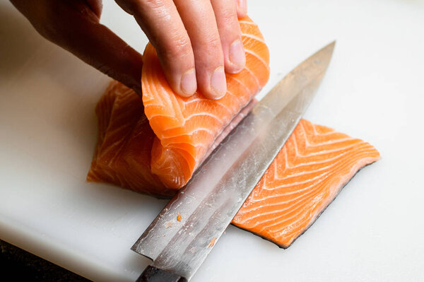 Профессиональный повар режет свежее филе лосося острым ножом на белой режущей поверхности. Вид сверху. Свежий лосось филе рыбы для приготовления сашими