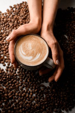 Sıcak taze kapuçino naer kavrulmuş kahve çekirdekli kadın eli. Üst Manzara. Kahve fincanı ve kadın eli kahve çekirdeğinin arka planında..
