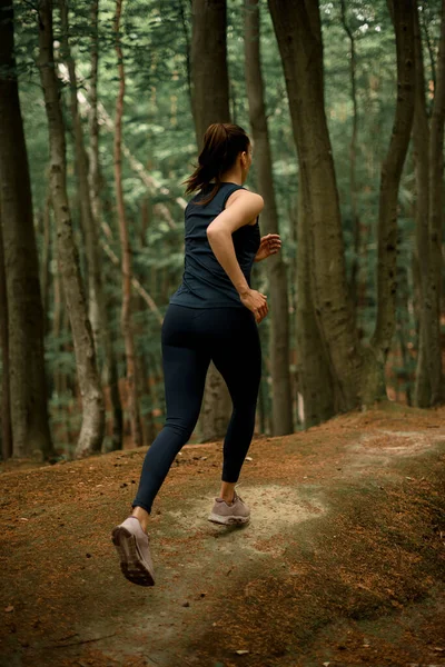Female with brown hair in pony tail running and jogging on hills in forest between tall deciduous trees. Sport and healthy lifestyle concept. Outdoor training