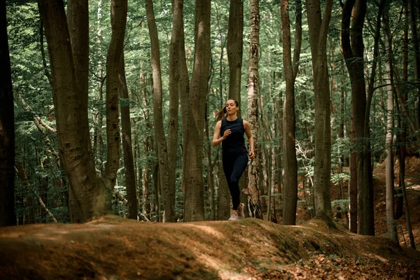 Woman with brown hair in pony tail running and jogging on hilly forest area between tall deciduous trees. Sport and healthy lifestyle concept. Outdoor training